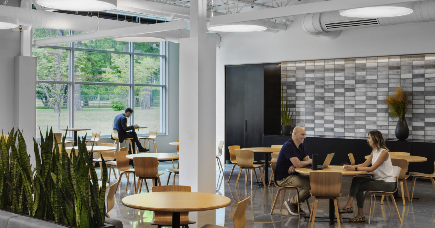 Modern office break room featuring natural light, with round wooden tables, beige chairs, and pendant lighting. A gray sofa flanks a green plant installation, enhancing the biophilic design. The exposed HVAC system adds an industrial touch above a geometrically tiled accent wall.