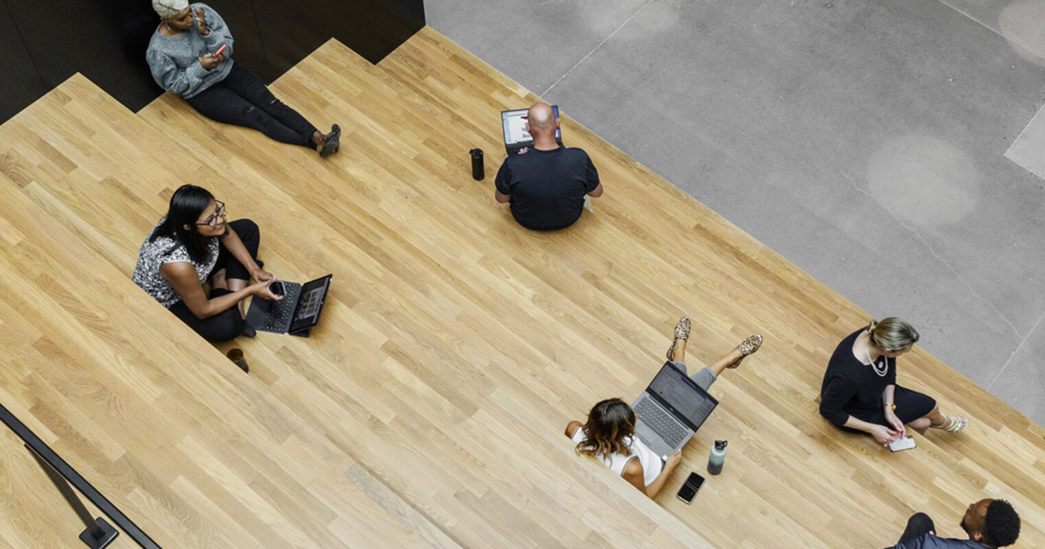 Overhead view of a modern office space featuring staggered wooden steps doubling as seating and work areas, with people engrossed in their laptops and mobile devices, highlighting flexible design and communal working environments.