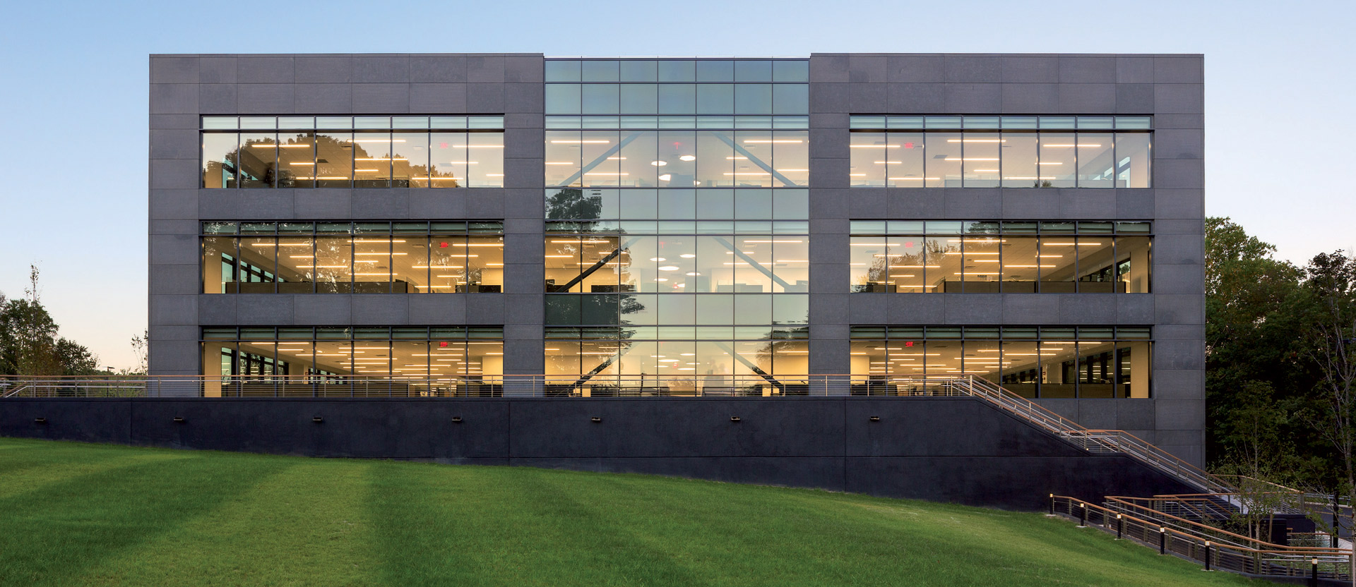 Modern office building at dusk with a fully glazed façade offering a glimpse of the multi-level, open-plan interior spaces, and warm artificial lighting that contrasts with the cool blue of the twilight sky. The building’s geometric symmetry and use of glass reflect contemporary architectural design principles.