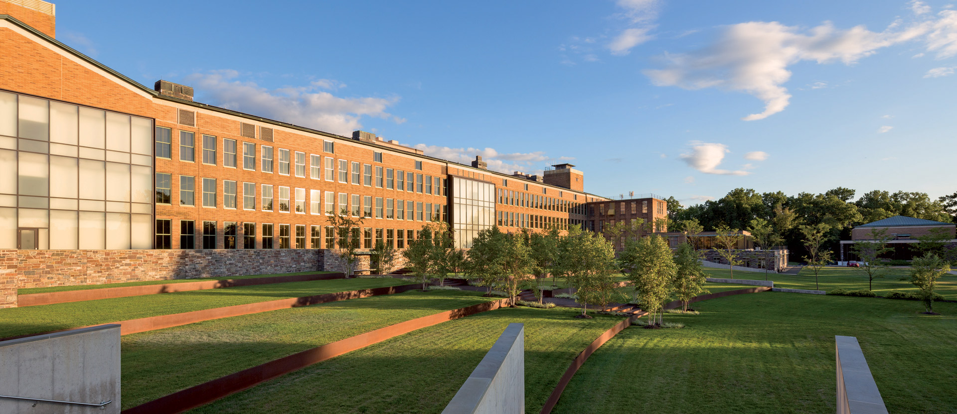 Modern educational facility with a blend of traditional brickwork and contemporary glass facade, landscaped with green lawns, geometric walkways, and young trees, under a clear blue sky.