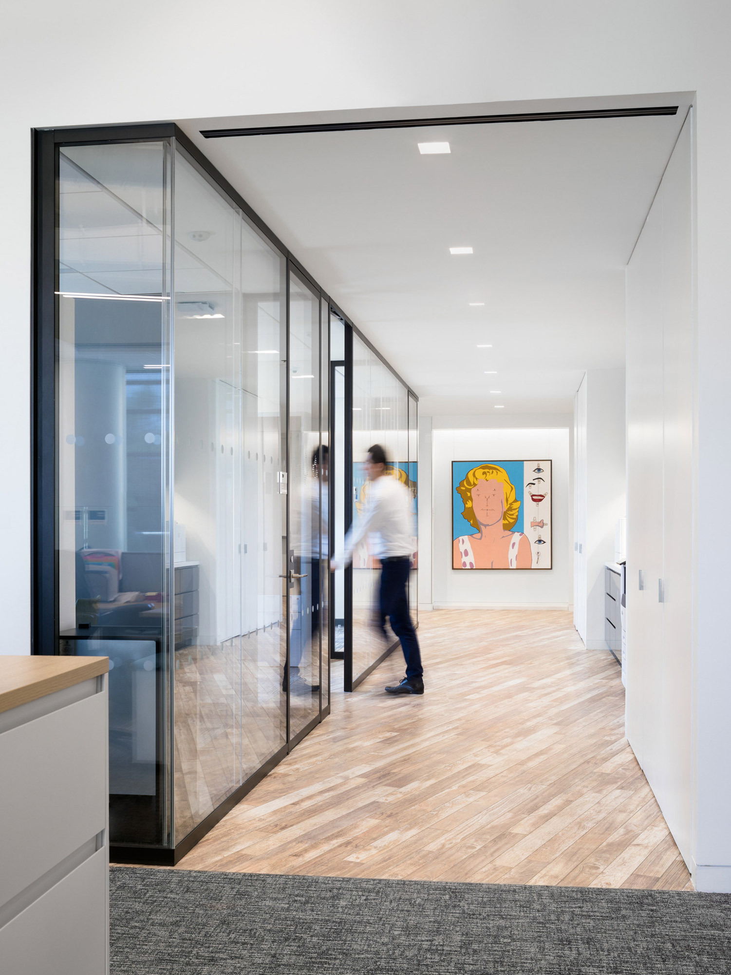 A modern office corridor with clear glass partition walls, light wooden flooring, and contemporary art on the right wall. A person walks past the semi-transparent meeting rooms, illustrating a blend of openness and privacy in the design.