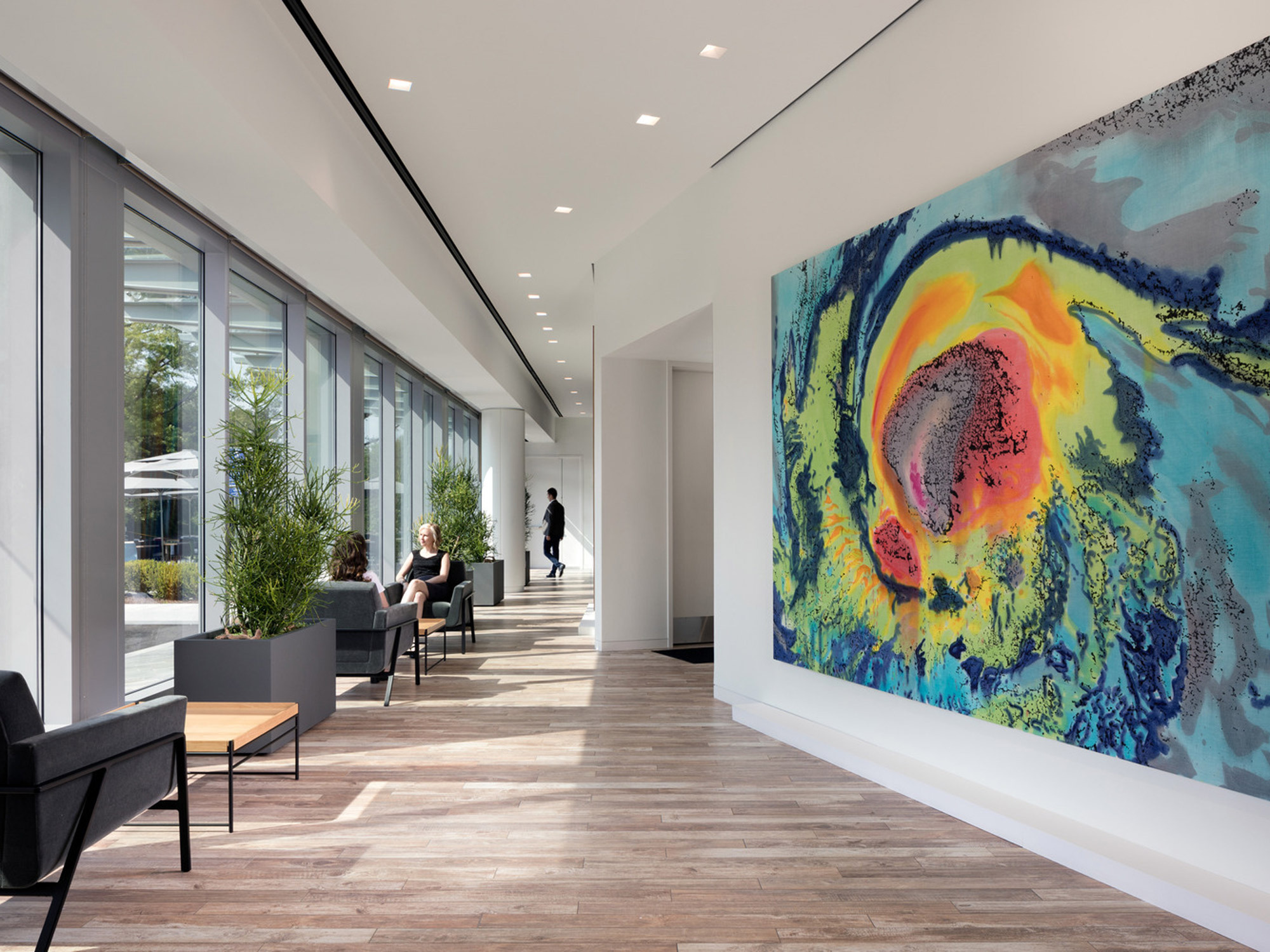 Modern hallway in a corporate building featuring sleek wooden floors, white walls, and expansive windows that infuse the space with natural light. A prominent, colorful abstract painting adds a vibrant focal point, while minimalist furniture provides functional seating areas for brief interactions.