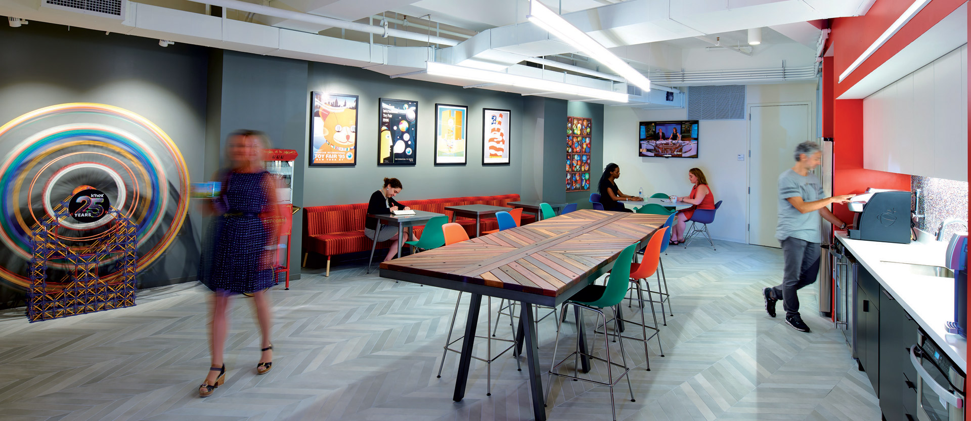 Modern office space featuring herringbone-patterned flooring, vibrant red and blue accents on walls and furniture, dynamic lighting, and contemporary artwork creating a lively, creative environment. Occupants engage in various activities within the collaborative, open-plan setting.