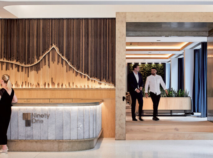 Modern office lobby with a curved wooden reception desk. Marble flooring transitions to a wooden passageway framed by elegant vertical wooden slats. Ambient lighting accentuates the smooth, natural materials and open, welcoming space.