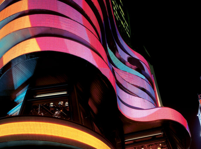 Night view of a dynamic building facade featuring undulating ribbons of LED screens, casting vibrant hues of red, blue, and yellow light. The structure's curves juxtapose against the surrounding urban signage, creating a modern, lively atmosphere.