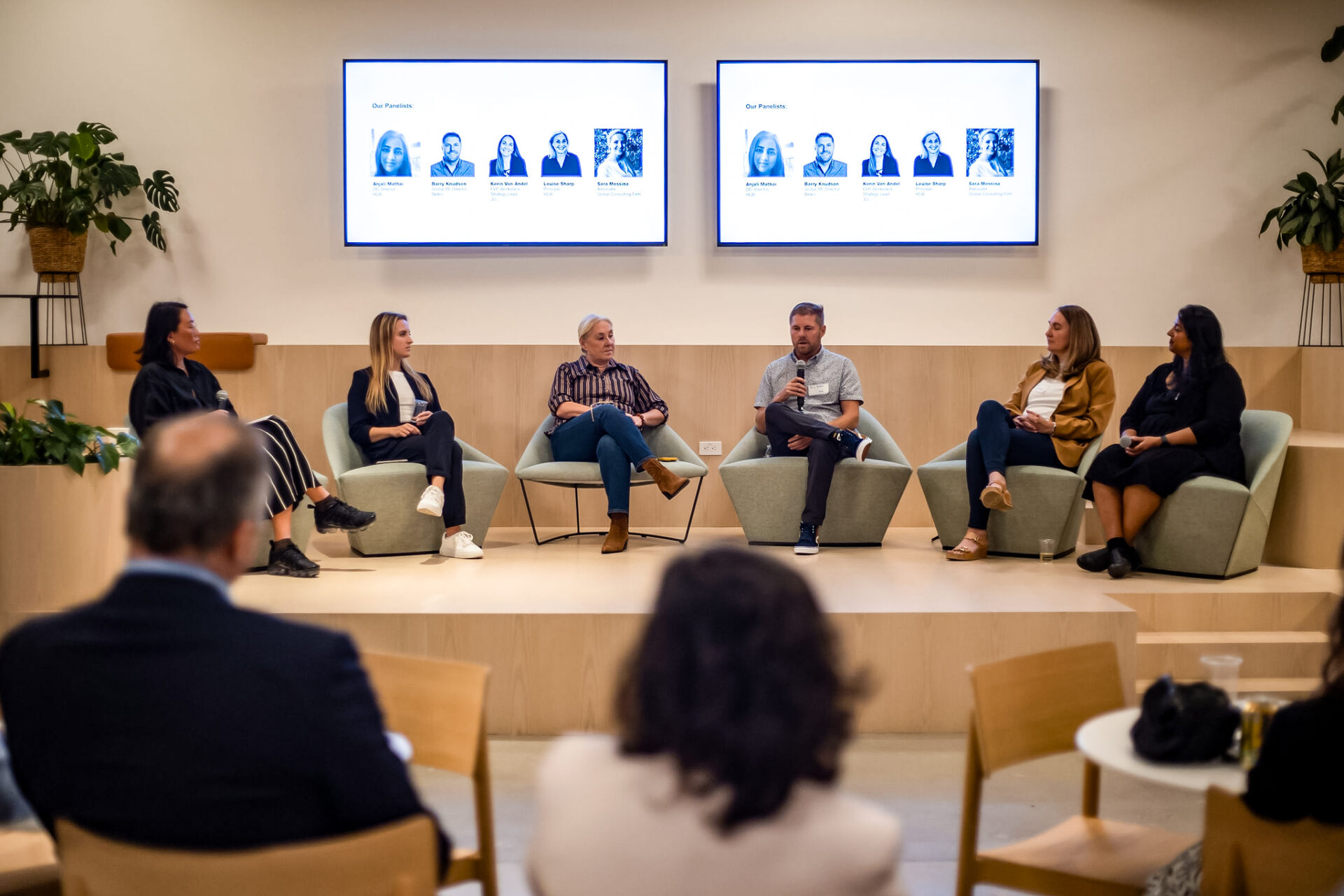Six individuals lead a panel discussion at the Belkin El Segundo workplace. The interior space is brightly lit with warm wood accents and biophilic elements like robust plantings.