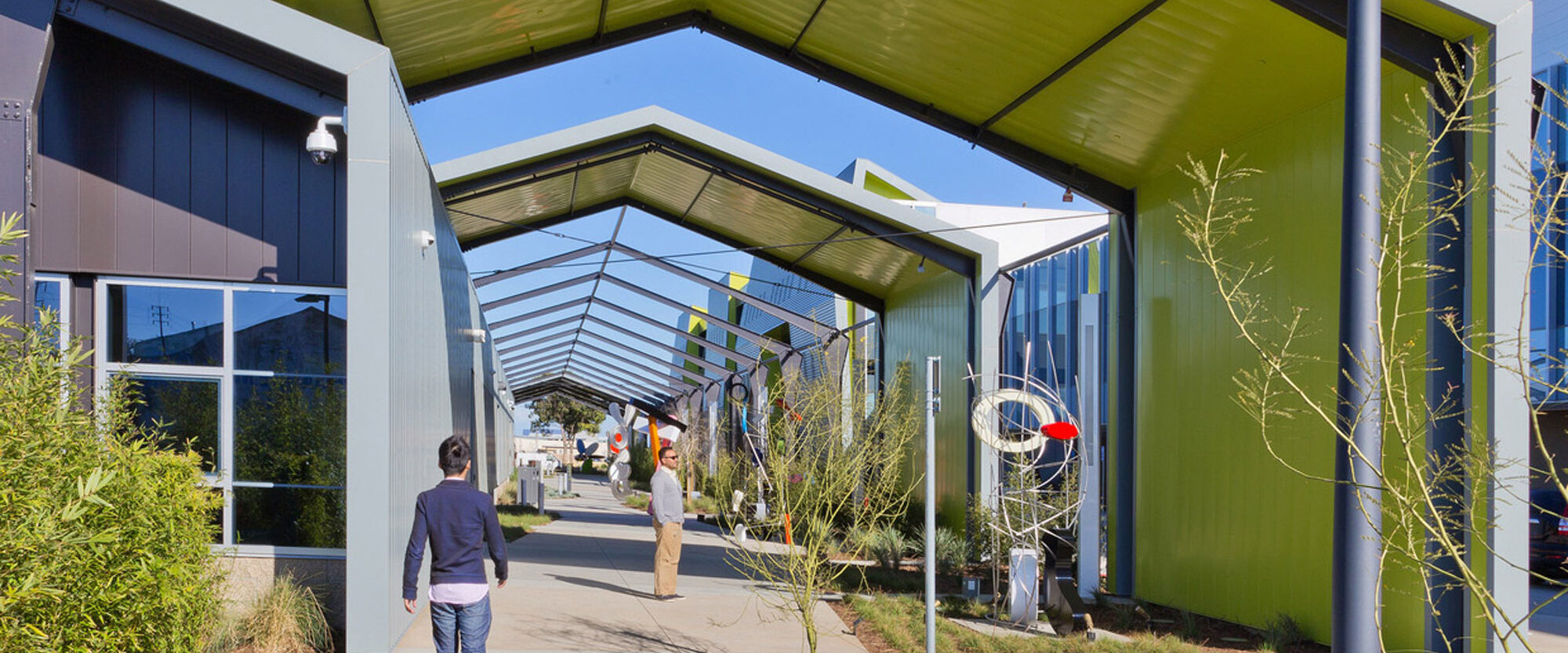 Modern commercial building with a striking green canopy stretching over the outdoor walkway, featuring geometric angles and contrasting blue sky, enhancing public space through architectural innovation.