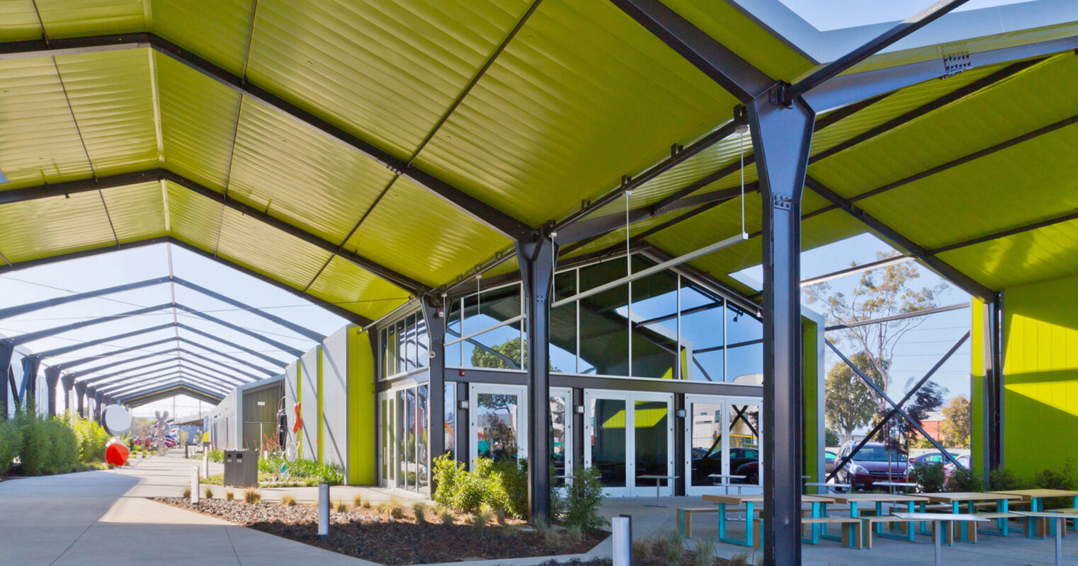 Bright, open-air walkway featuring a row of geometric, green canopies supported by steel columns, integrated into a modern commercial structure with large glass panels, reflecting a fusion of functional and eco-friendly design.
