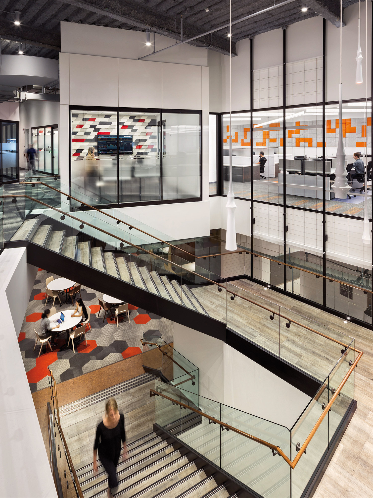 Modern office interior with an open floor plan features a floating glass staircase connecting two levels, accented by sleek metal handrails. The design incorporates a mix of neutral toned floor tiles and vibrant orange wall accents, optimizing the space for both collaborative and independent work areas.
