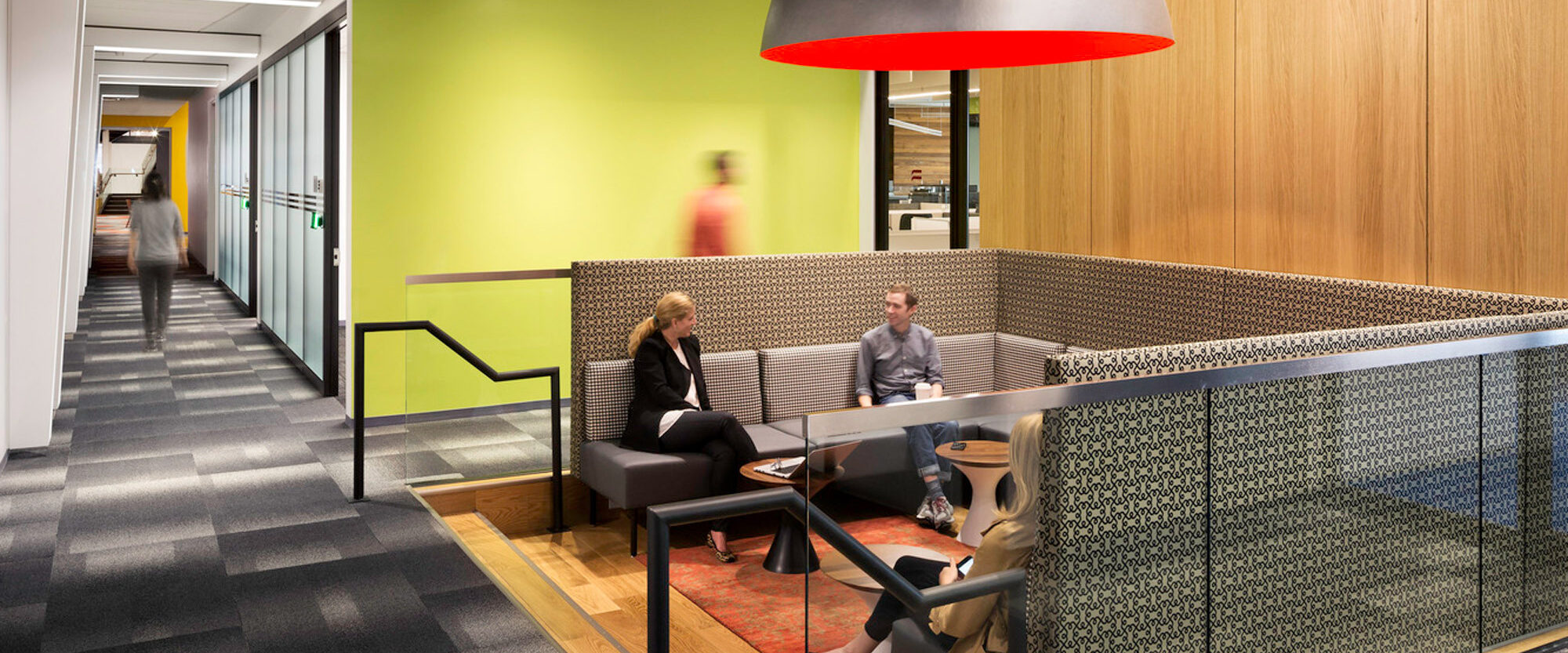 Modern office breakout space with geometric patterned booth seating, a large black and red pendant light, vibrant green walls, and wood paneling, designed for casual collaboration, bordered by a carpeted hallway leading to private work areas.