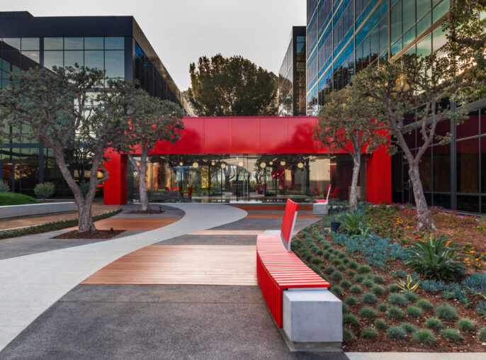 Landscape architecture integrates with modern corporate building design featuring a vibrant red awning, matching linear benches, and a meandering pathway amidst drought-tolerant plantings.