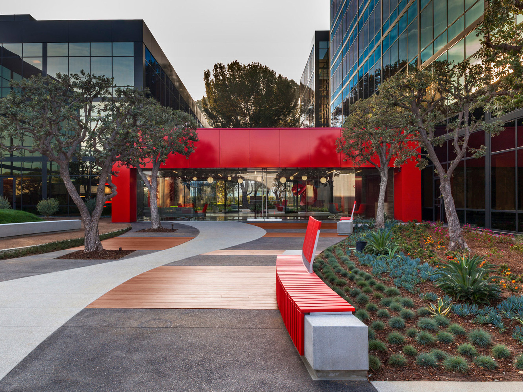 Landscape architecture integrates with modern corporate building design featuring a vibrant red awning, matching linear benches, and a meandering pathway amidst drought-tolerant plantings.