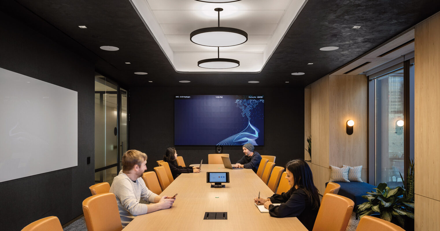 Modern corporate meeting room with a sleek design, featuring a long table with individuals engaged in a discussion, ambient lighting from circular fixtures above, and large screens for presentations on the wall.