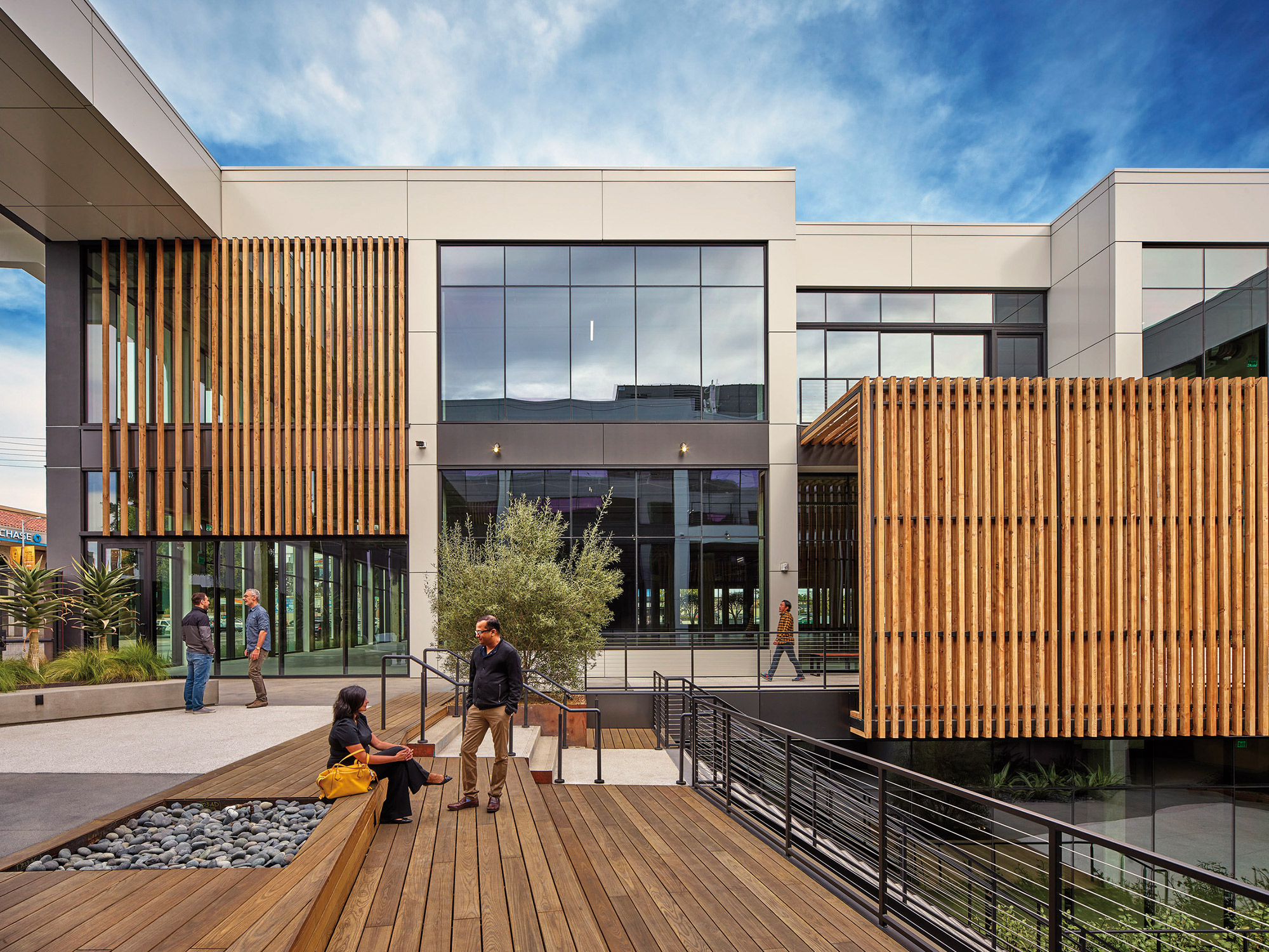 Modern office building with an outdoor seating area featuring wood slat accents, where people engage in casual conversation.