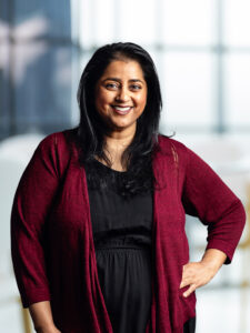 The photograph captures a woman with long black hair and a friendly smile, wearing a black dress and a burgundy cardigan. The backdrop is bright and blurred, featuring vertical lines and hints of blue, suggesting a modern office environment with large windows.