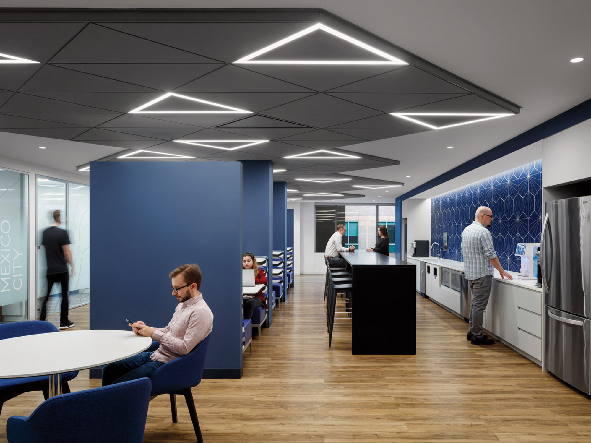 Modern office kitchen and break area featuring bold blue partitions, geometric ceiling lights, and wood flooring. White countertops and stainless steel appliances complement the space where employees are casually interacting.