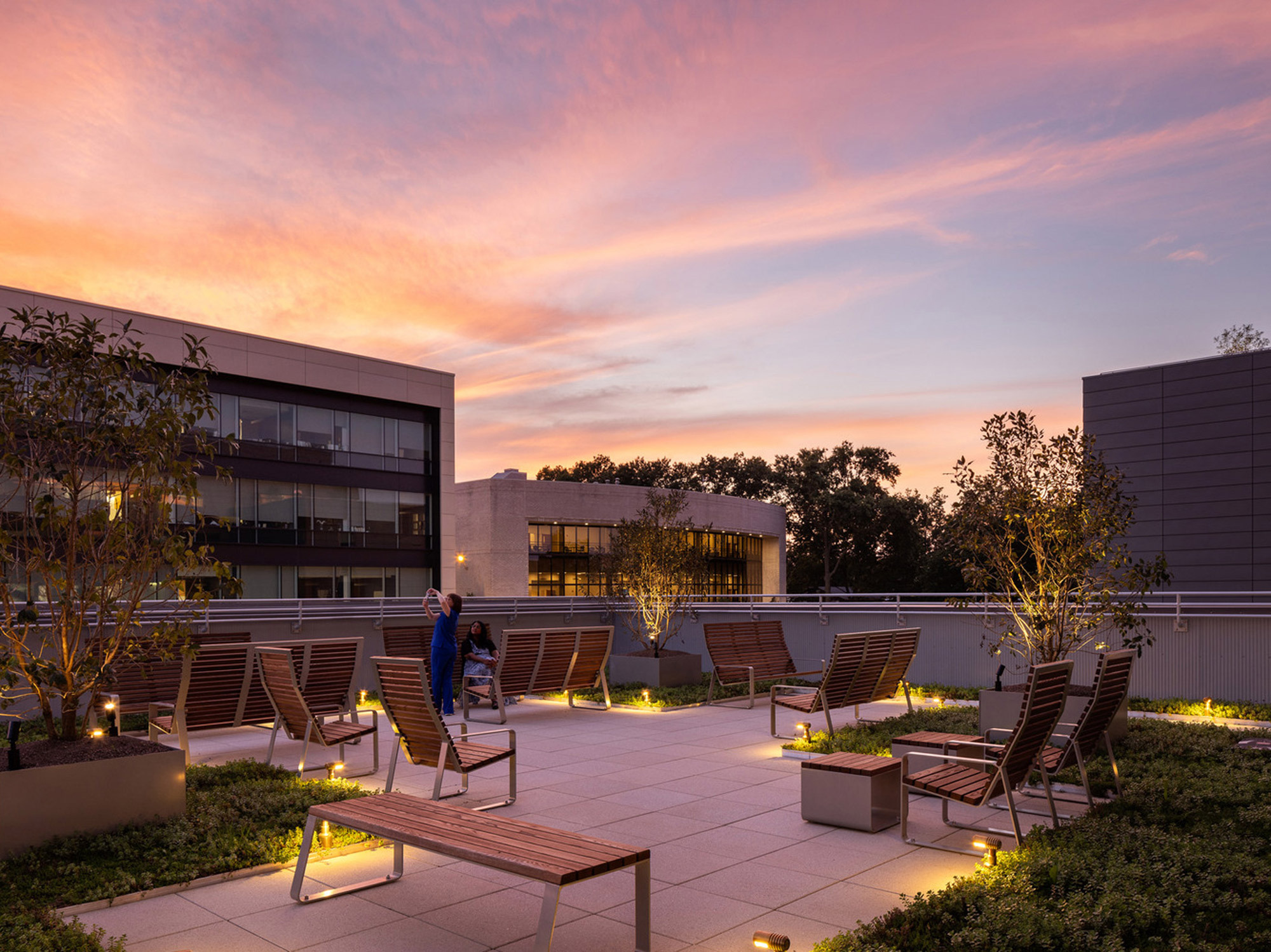 Rooftop terrace at dusk with integrated wooden seating and planters, under-floor lighting, against a backdrop of modern architecture and a vibrant sunset sky.