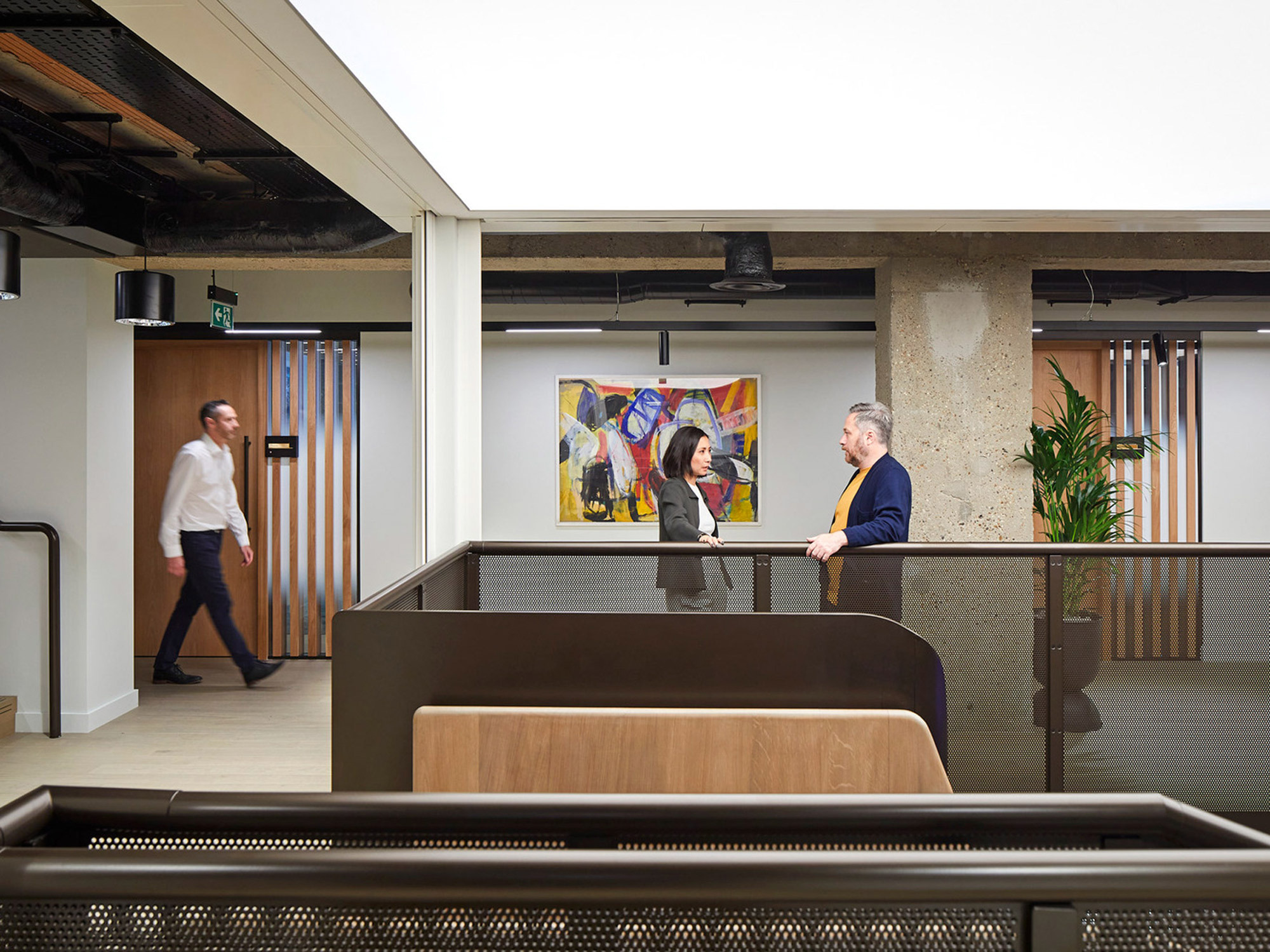 Open-plan office space featuring industrial design elements with exposed ceiling beams and concrete pillars. Two professionals engage in conversation near a glass partition with vibrant abstract art, while another walks by, emphasizing the dynamic work environment. Minimalist wood and black metal reception desk foreground.