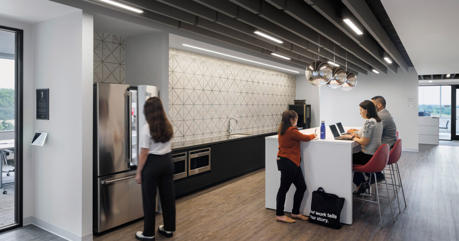 Modern office kitchen featuring stainless steel appliances, white cabinetry, and geometric backsplash. Pendant lighting illuminates a central island where employees converse casually. Monochrome color palette with natural light accentuating clean lines and minimalist design.