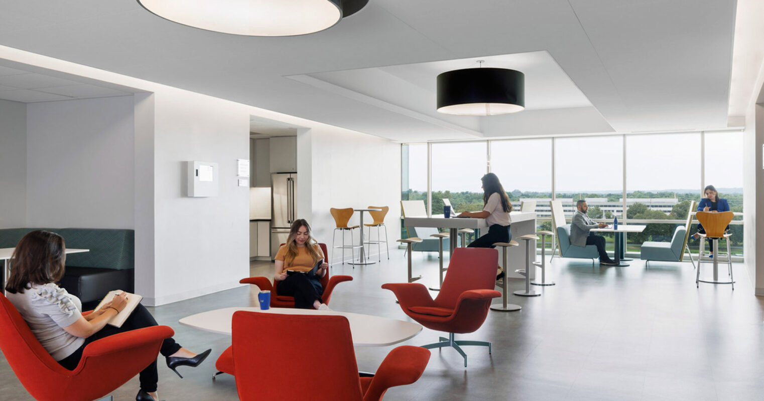 Open-plan office lounge with red armchairs centered around low coffee tables. The space is illuminated by oversized black pendant lighting and expansive windows providing abundant natural light. Neutral floors and walls emphasize the bold furniture color. Individuals are seated variously, engaging in work and conversation.