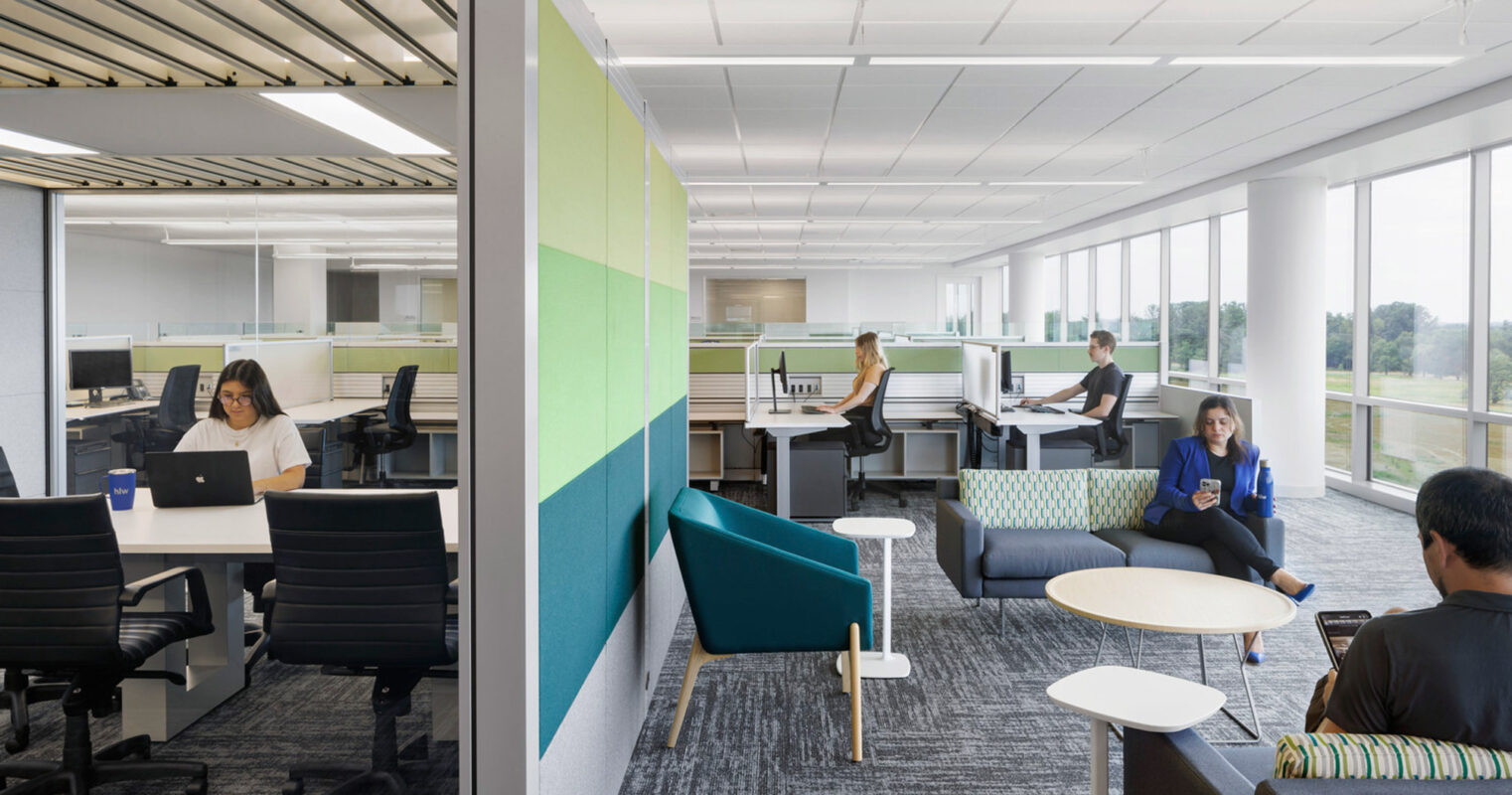 Open-plan office space featuring modular workstations with ergonomic chairs, individual privacy panels, and ambient natural lighting from floor-to-ceiling windows. A casual seating area introduces vibrant upholstery, complementing the space's contemporary, collaborative atmosphere.