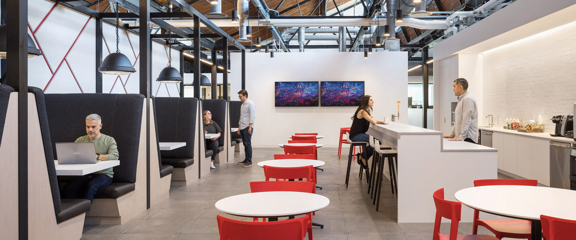 Modern open-plan office space featuring a high ceiling with exposed ductwork, sleek red tables and chairs, private cubicles with gray acoustic panels, and a centralized kitchen area with bar seating, under warm pendant lighting.