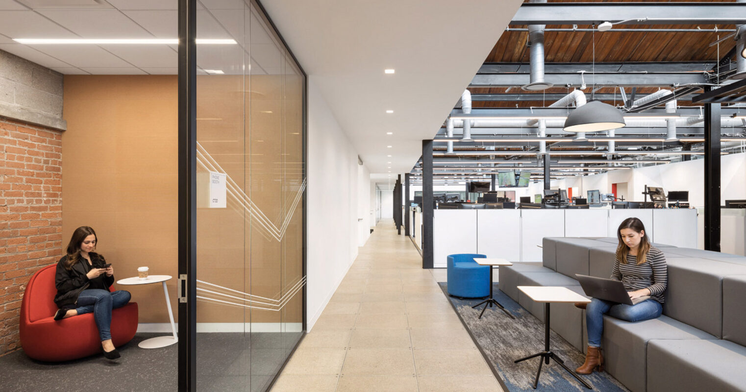 Modern office space with exposed ceiling beams, featuring an open plan seating arrangement with individual workstations. A communal lounge area includes colorful, contemporary furnishings and glass partitions, creating a blend of private and collaborative environments.