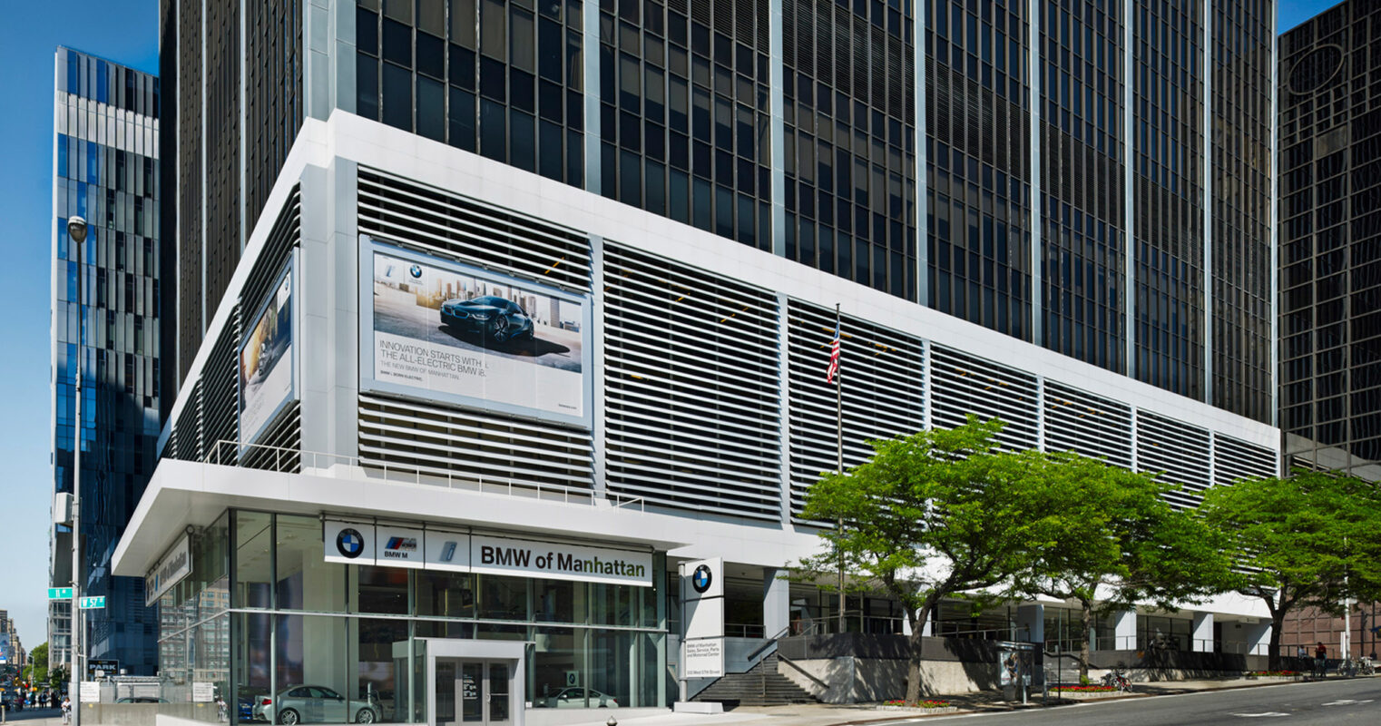 Modern car dealership exterior with prominent white horizontal louvers above glass storefront, framed by minimalist steel columns, set against a backdrop of a bustling metropolitan street and towering skyscrapers.