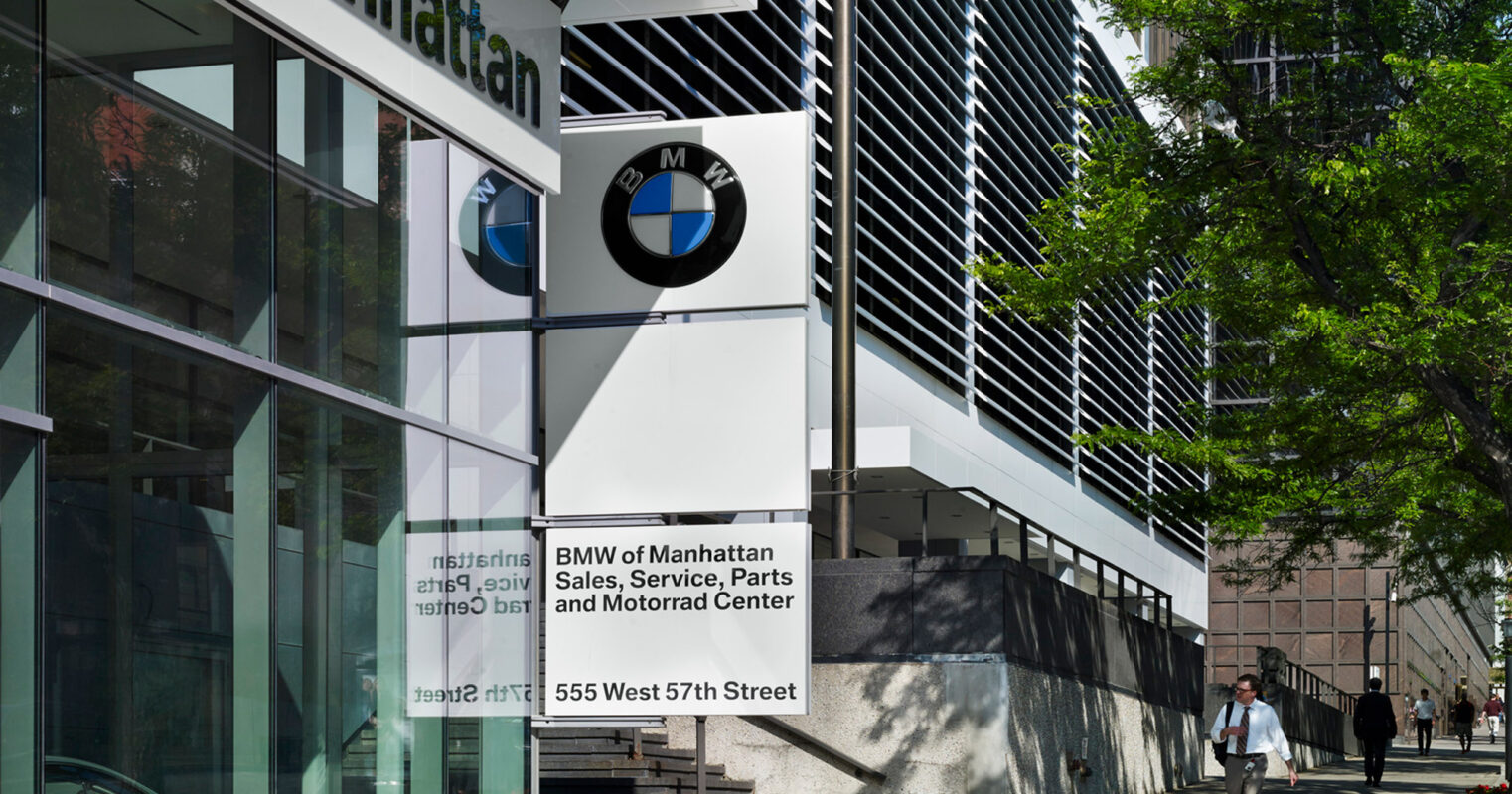 Exterior view of BMW of Manhattan building showcasing modern architecture with white angled louvers, large windows, and prominent signage, nestled in an urban setting.