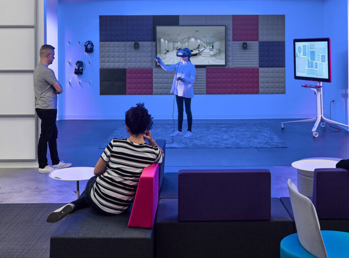 Modern office break room featuring a vibrant color scheme with purple and blue modular seating, a paneled red and gray acoustic wall, and an interactive whiteboard for presentations, fostering a collaborative and creative work environment.