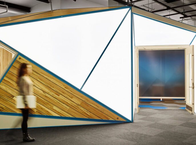 Modern office space featuring angular meeting pods with wooden slats and teal accents. Sleek, exposed ceiling fixtures add an industrial touch above the dynamic workspace layout.