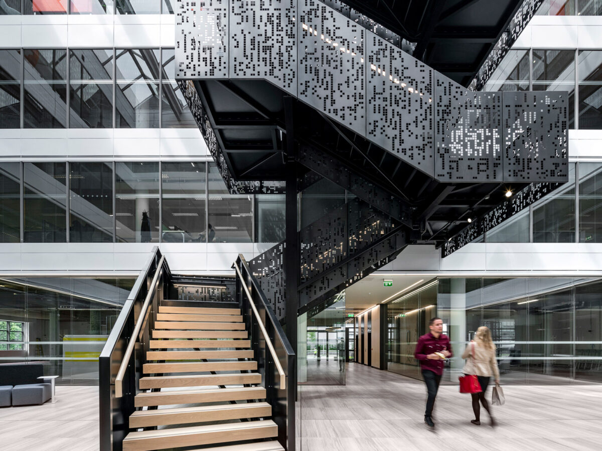 Modern office atrium with a geometric black staircase featuring perforated patterns, connecting multiple levels. Glass balustrades and open-plan layout create a sense of transparency and flow, juxtaposed by bold, angular lines of the central stairway.