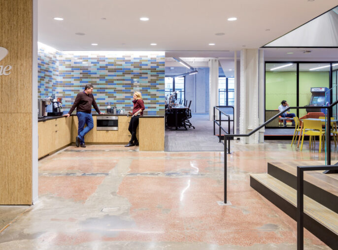 Spacious commercial lobby with mix of natural and industrial elements, featuring a terrazzo floor, wood-paneled reception desk with the Capital One logo, mosaic tile wall art, and a staircase leading to an upper level with occupants in motion, reflecting dynamic workplace design.