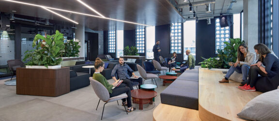 Spacious modern office break area featuring a mix of seating options, from plush sofas to wooden benches, under a distinctive lighting scheme with a dynamic, wavy LED track. A communal kitchen is anchored by potted plants, promoting an inviting and comfortable collaborative workspace.