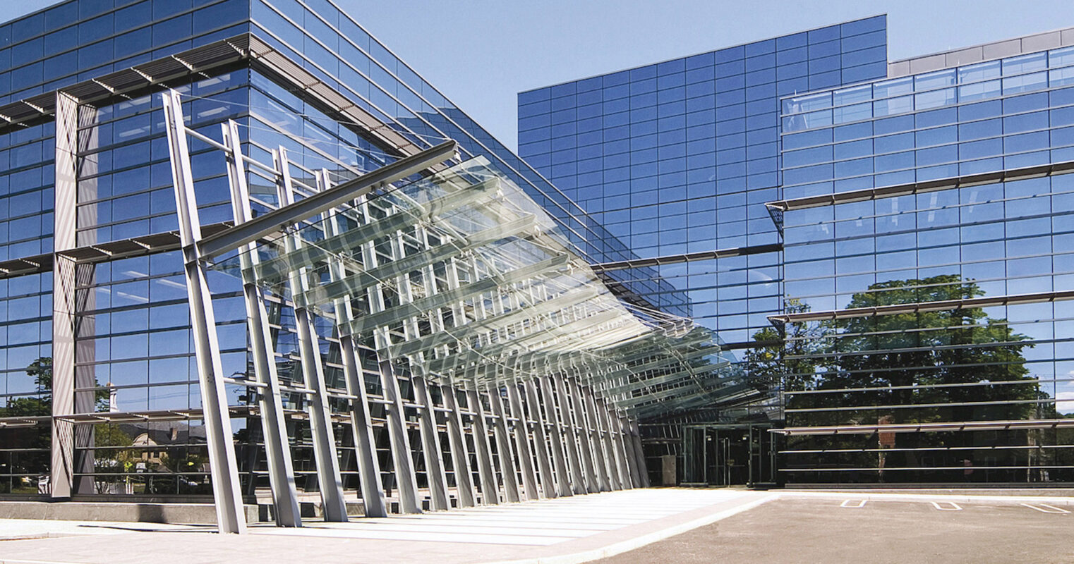 Modern corporate building entrance featuring a prominent glass structure, reflective façade, and strong geometric lines that create a striking visual symmetry against the blue sky.
