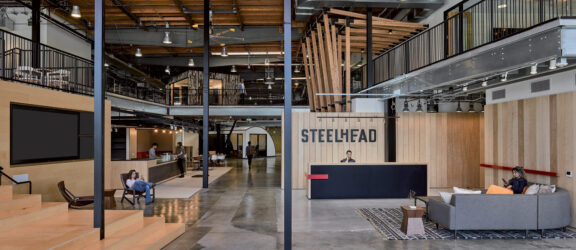 Spacious industrial-style office interior featuring exposed wooden beams, HVAC ductwork, and pendant lighting. The open-plan layout includes a mezzanine level, modern furnishings, and a muted color palette that accentuates the natural wood and metalwork. The Steelhead branded reception area anchors the design.