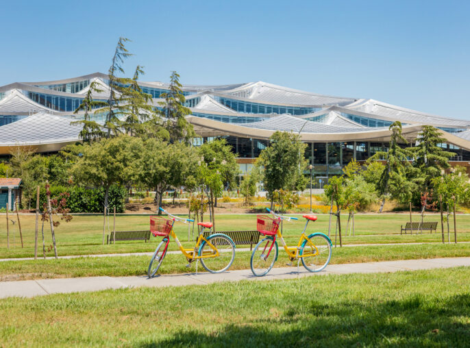Lush green lawn fronts a modern architectural marvel with undulating glass facade and white supports, flanked by vibrant yellow bicycles in sunny park-like setting.