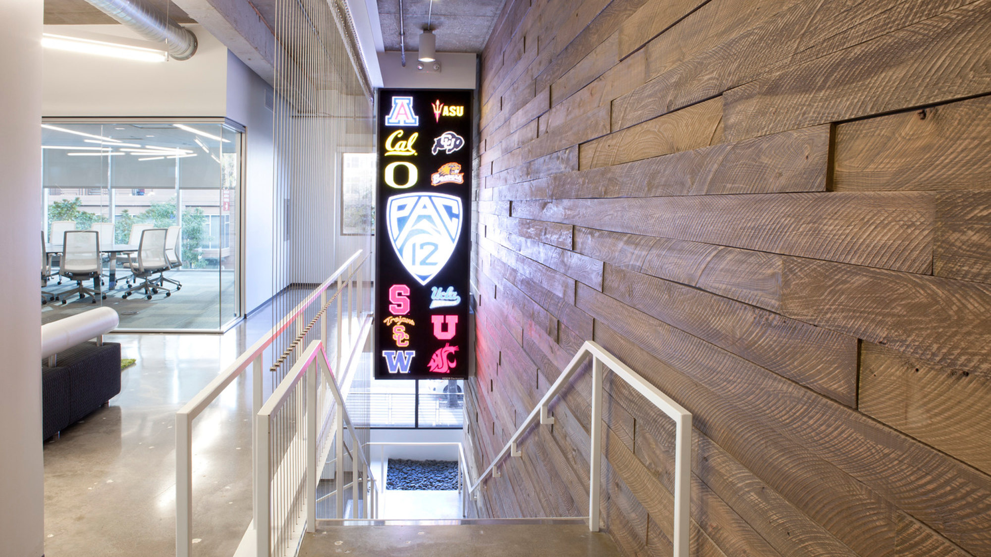Contemporary office stairwell with reclaimed wood wall cladding and collection of vibrant college sports team neon signs, exhibiting a blend of rustic textures and modern collegiate pride, leading to an airy workspace with glass partitions.