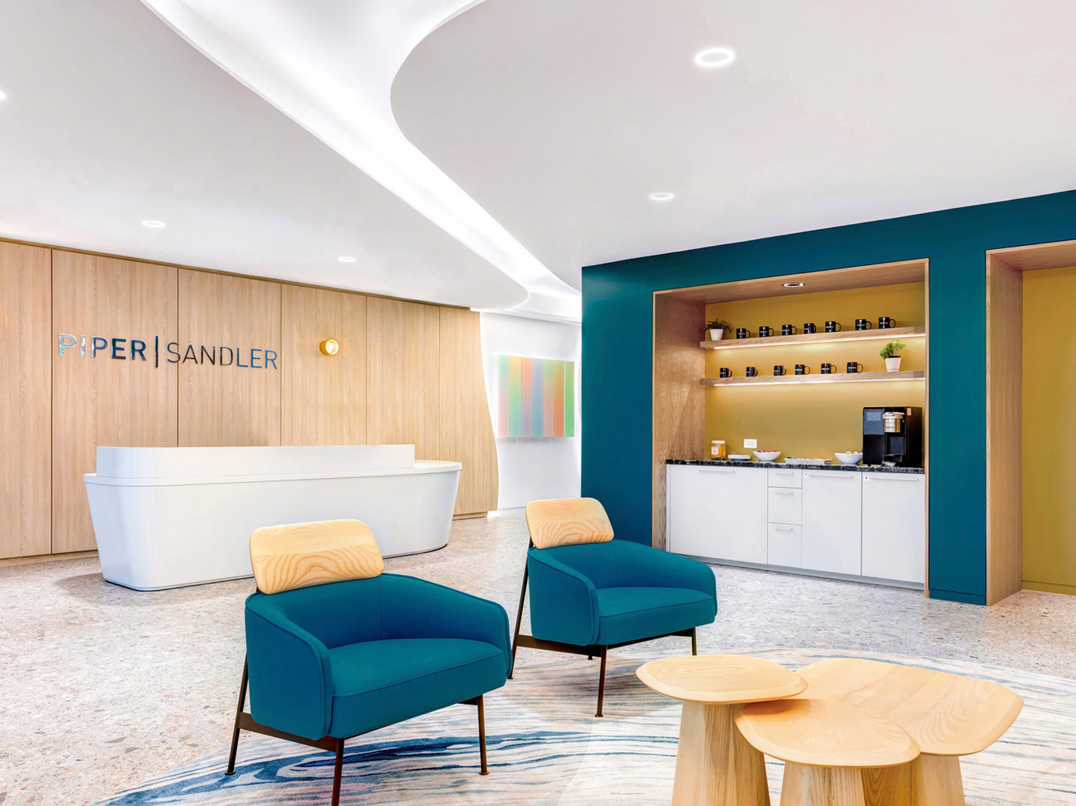 Modern reception area with curved white desk, accented by teal armchairs and wooden stools. A blue alcove with shelving hosts refreshments, set against a warm wooden backdrop with the company’s signage. Ambient lighting complements the room's clean, inviting aesthetic.