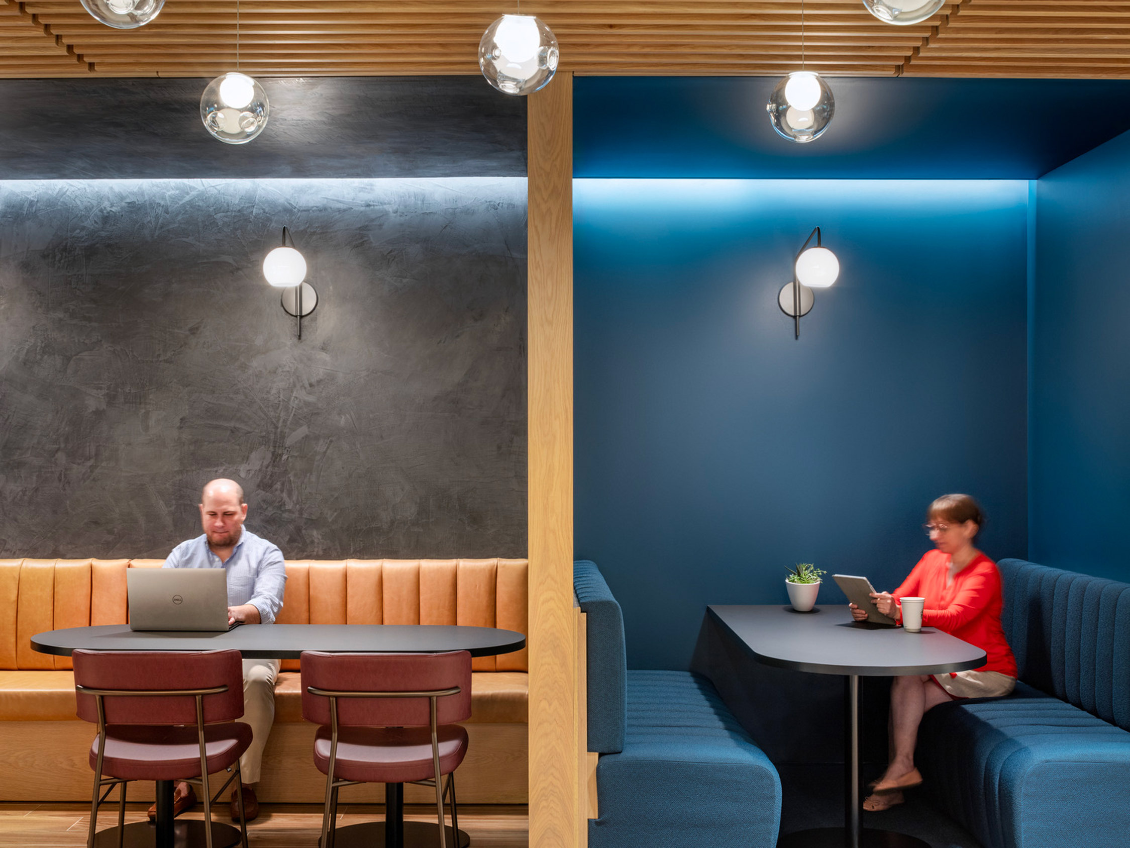 Modern office breakout area featuring cobalt blue walls, warm wood accents, and integrated lighting. Mustard-toned, tufted seating complements the space while professionals engage in focused work at individual tables.