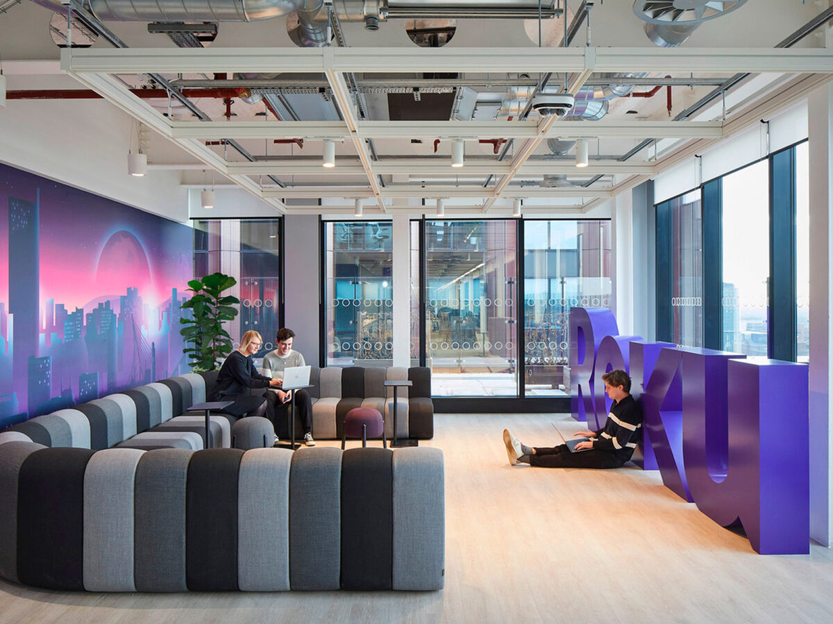 Modern office lounge with modular gray seating arrangements, vibrant purple partitions, industrial ceiling with exposed ductwork, and cityscape mural providing a dynamic backdrop. Natural light streams in through floor-to-ceiling windows, and employees engage casually around a central coffee table.