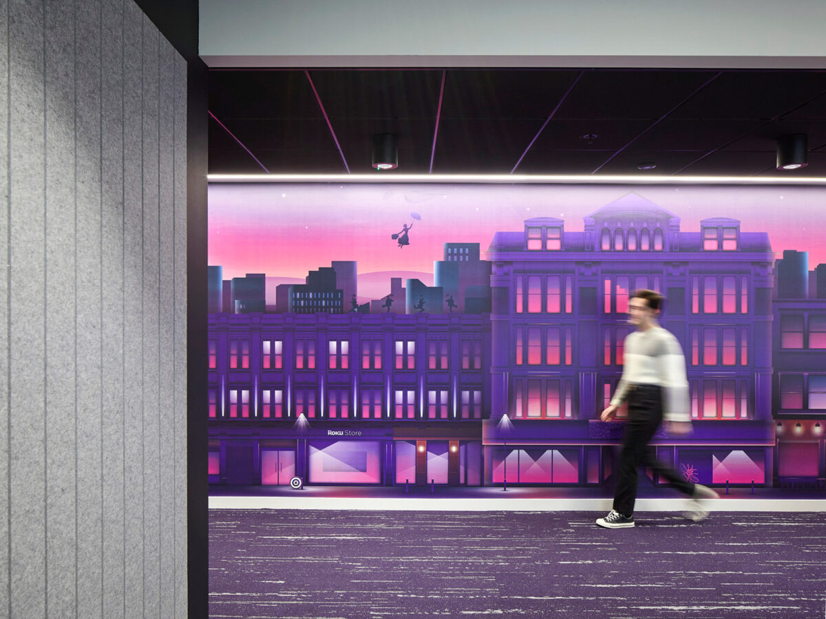 Wall mural in office corridor featuring a stylized cityscape at dusk with dynamic lighting, blending real architectural elements with graphic illustration. A silhouetted figure walks by, adding life to the scene.