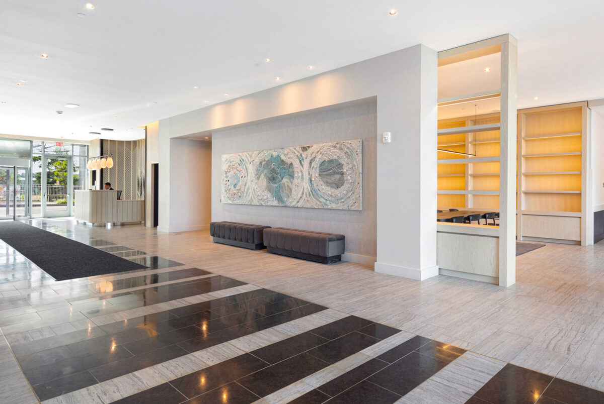 Spacious lobby with polished tile flooring in a checkerboard pattern, flanked by minimalist grey columns. The focal point is an expansive textured art piece in cool tones above an elongated, tufted bench. Symmetrical shelving with illuminated niches complements the sleek and modern aesthetics.