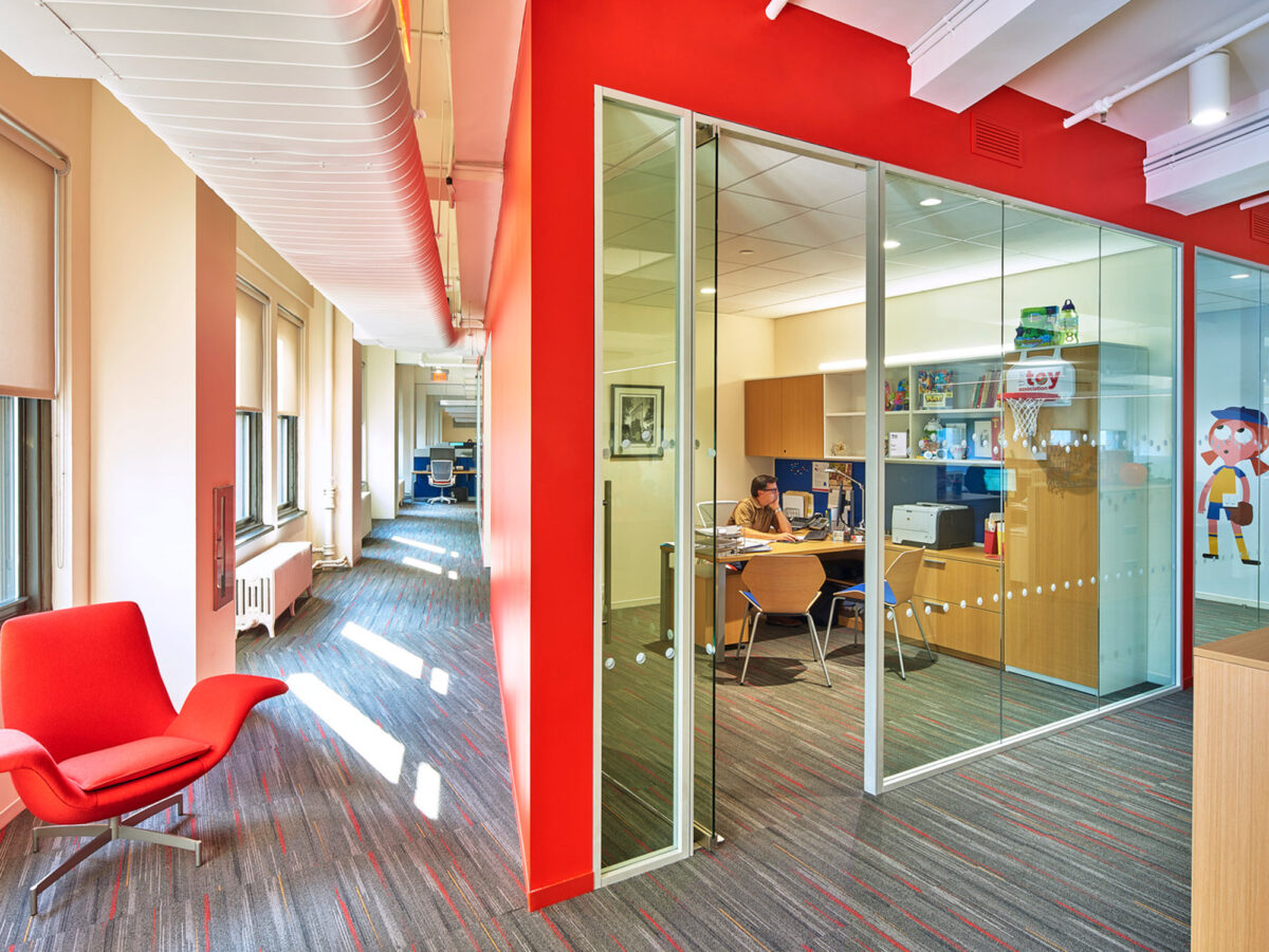 Contemporary office space featuring a bold red chair, walls in contrasting red and white, and glass partitions. Exposed white ceiling ducts provide an industrial feel, while the transparent office walls create an open atmosphere. A person is at work at a desk inside the glass-walled room.