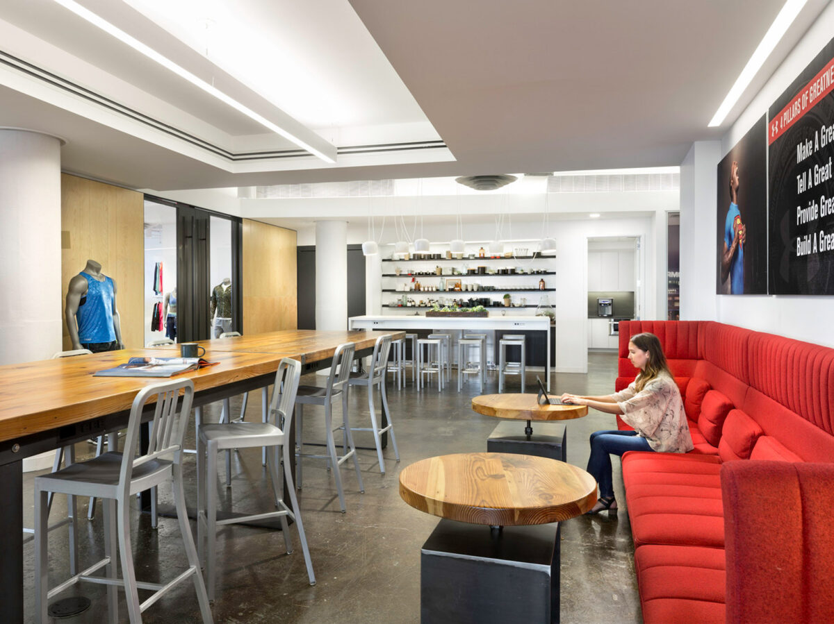 Contemporary office break room featuring sleek white countertops, a red upholstered banquette, industrial-style stools, and circular wooden tables. Integrated lighting accents the modern ambiance while a fashionable clothing display adds a creative visual element.