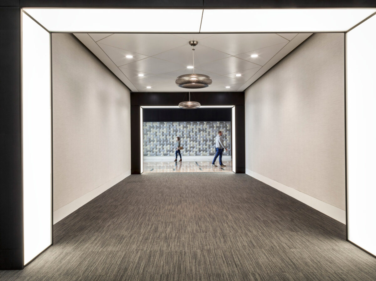 Sleek, modern corridor with recessed lighting highlighting textured walls and an artistic, patterned backdrop at the end. Two individuals in professional attire walk along the carpeted floor, conveying a dynamic professional environment. The interior embraces a monochromatic palette punctuated by the intricate feature wall.