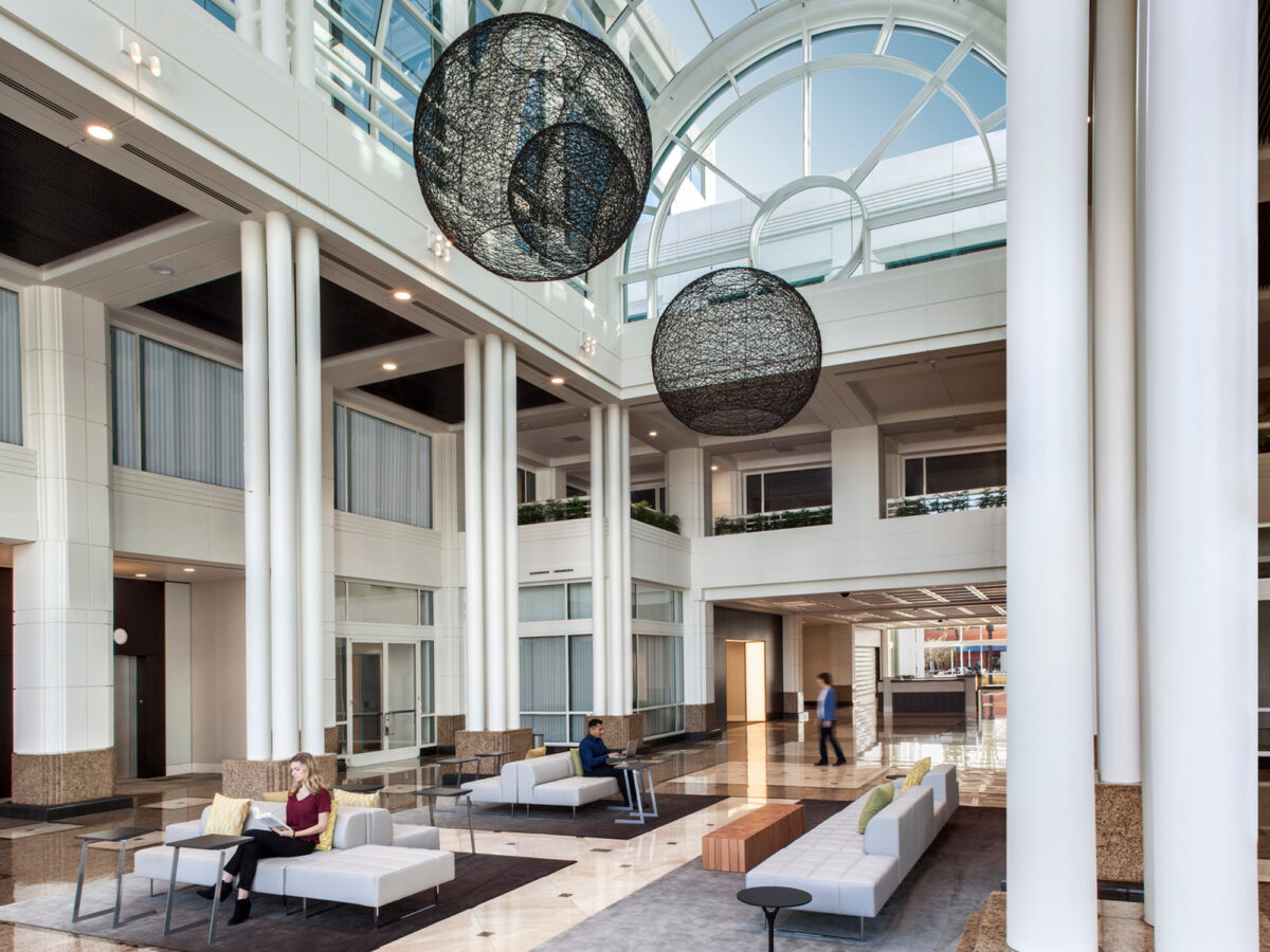 Spacious modern lobby featuring high ceilings with two large spherical mesh pendant lights, white modular seating, clerestory windows, and a mix of natural stone pillars and wood paneling, conveying a luxurious, airy atmosphere.
