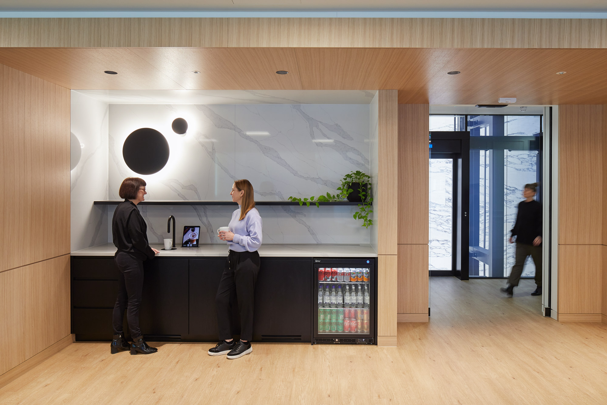 A modern office kitchenette, elegantly designed with a blend of natural wood and marble finishes. Two professionals are engaged in conversation by the countertop, which is set against a striking marble backsplash adorned with abstract black and white patterns. The space is accentuated by minimalist black cabinetry and a wooden archway framing the entrance. Soft lighting and a small plant add a welcoming touch, while a beverage fridge provides functionality, enhancing the comfort and usability of the area.