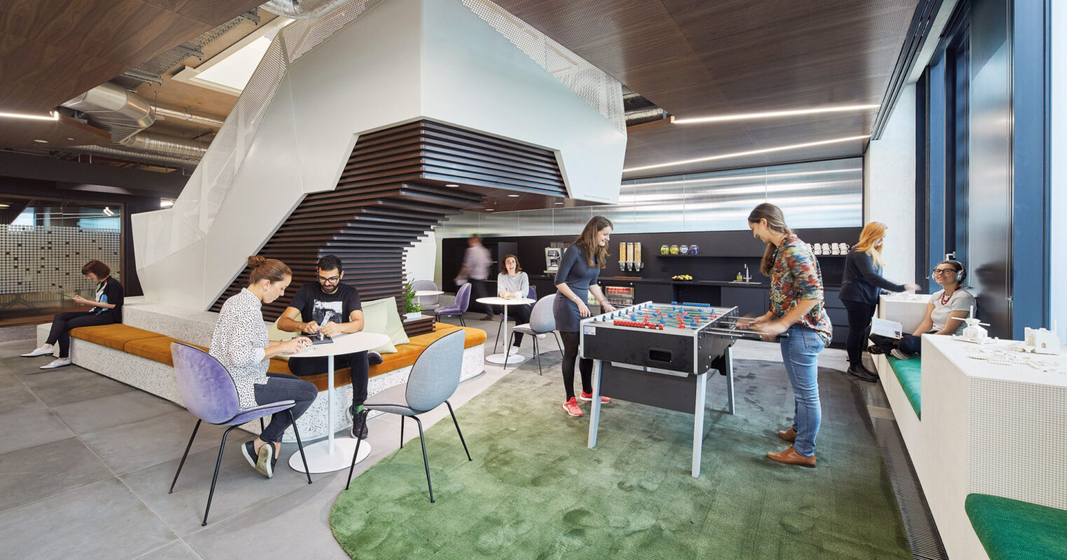 Modern open-plan office space with a variety of seating arrangements featuring eclectic furniture. Employees engage casually around a foosball table. Overhead, exposed ductwork complements the industrial-chic aesthetic, while vibrant green carpeting adds a pop of color, aiding in zone delineation.