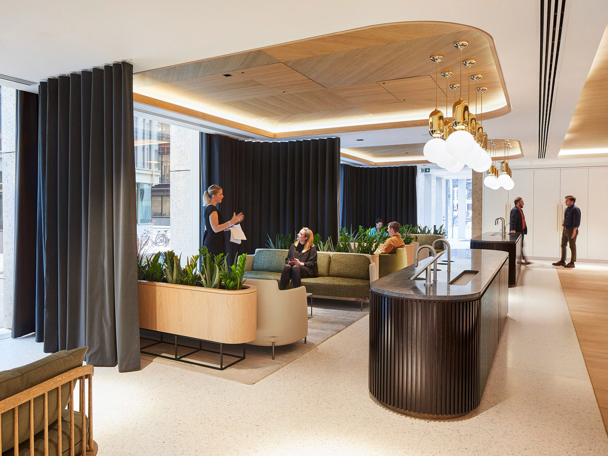 Modern office lobby with a warm, welcoming atmosphere, featuring curved wooden reception desk, elegant suspended light fixtures, and vertical drapery creating private nooks. Rich plant arrangements add a natural touch, while people converse and work, emphasizing the space's functional design.